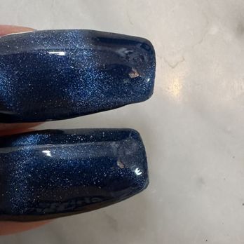 V Nails - The image displays two fingers with freshly painted dark blue nail polish that contains glitter particles, giving a sparkly appearance. The nails are well-manicured and glossy, with a smooth finish. The background appears to be a light-colored surface, possibly a table or countertop, indicating a setting related to nail care or beauty services. - Nail Salons Near Me - Avon, Indiana