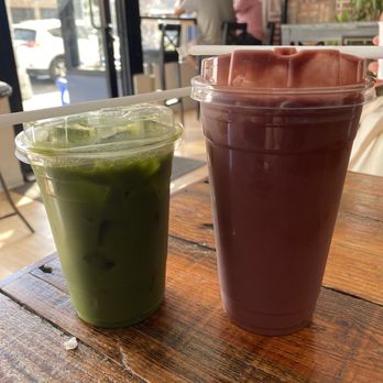 IC Brooklyn Cafe - Iced Matcha Green Tea Latte and Brazilian Sunrise Smoothie - Two iced drinks, one green matcha latte and one dark chocolate beverage, are on a wooden table inside a cafe. The cafe has large windows allowing natural light, with two people sitting at a table in the background. The interior features brick walls and potted plants, creating a cozy atmosphere. - Bagels Near Me - Brooklyn, New York