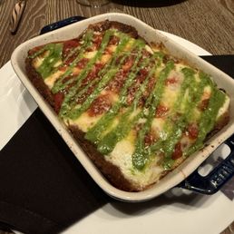 Eggplant Parm - A baked eggplant parmesan dish in a rectangular ceramic baking dish features a layer of melted cheese, tomato sauce, and green pesto drizzle on top. The dish is placed on a white plate with a black napkin underneath, on a wooden table. In the background, there is a plate with a different dish and a glass of wine, indicating a restaurant setting.
