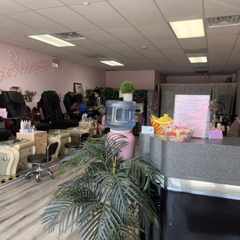 Super Star Nails - Interior - The image shows the interior of a licensed nail salon with a row of black and beige pedicure chairs along the left side, each with small tables holding nail supplies. The ceiling has fluorescent lighting, and the walls are painted light pink with decorative wall art. In the background, clients are seated receiving nail services, with staff attending to them. The reception area on the right includes a counter with a potted plant, a bowl of candies, and a staff member wearing a mask. The space appears clean and organized, with a water dispenser and a television mounted on the wall. - Nail Salons Near Me - Frankfort, Illinois