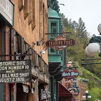 Collette Tours - Where Wild Bill Hickok was shot, Deadwood, SD - A street scene in Deadwood, South Dakota, displays vintage storefronts and signs, including the Deadwood Trading Post, Sick Boy Motorcycles, and a historic site marker indicating where Wild Bill Hickok was shot in 1876. The street is lined with old-fashioned lamp posts and a hillside with trees in the background, capturing a historic and nostalgic atmosphere. - Tours Near Me - Springfield, Massachusetts