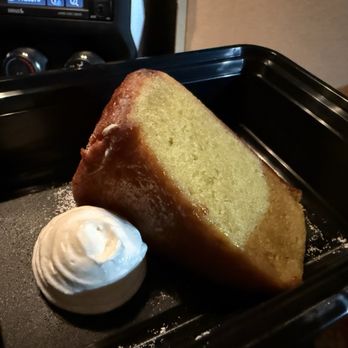 Penelope - Olive Oil Cake - A slice of olive oil cake with a golden-brown crust and moist, yellow interior is placed on a black tray. Next to the cake, there is a dollop of whipped cream. The background shows part of a kitchen or restaurant setting with a stove or oven controls visible. The presentation suggests a dessert served at a restaurant or cafe. - Pizza Near Me - Tucson, Arizona