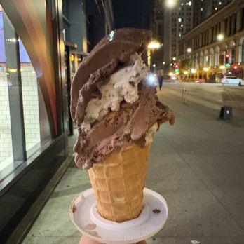 The Original Rainbow Cone - Chocolate Obsession - A person is holding a waffle cone filled with multiple scoops of chocolate and vanilla ice cream. The photo is taken outdoors at night, with city buildings, streetlights, and traffic visible in the background. The scene suggests an urban setting with a lively nighttime atmosphere. - Ice Cream & Frozen Yogurt Near Me - Chicago, Illinois