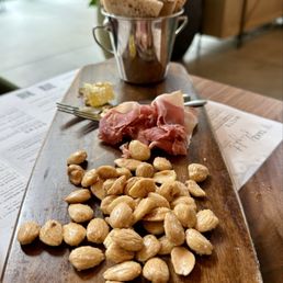 Charcuterie - The image features a wooden serving platter with a selection of food items including slices of prosciutto, roasted nuts, a small portion of honey, and a metal bucket containing slices of bread. The platter is placed on a table with menus visible underneath, and the background shows a bright, indoor setting with plants and natural light.