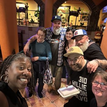Los Potrillos - Inside - A group of eight people, including men and women of diverse backgrounds, are gathered in a restaurant with warm orange walls and arched architectural features. They are smiling and appear to be enjoying each other's company. Some are wearing casual clothing, including hats and glasses, and one person is holding a takeout container. The background shows a window with plants and a television screen, indicating an indoor dining setting. - Mexican Near Me - Cincinnati, Ohio
