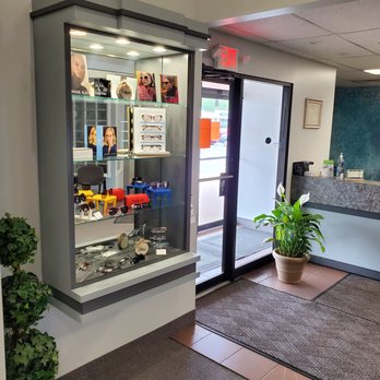 Vision Care Coventry - The image shows a glass display case containing various eyeglasses, sunglasses, and related accessories, situated inside an optometry office. The entrance door is visible nearby, with a potted plant on the floor and a reception desk in the background. The setting includes indoor lighting, a carpeted floor, and a wall with the partial word 'VISION' visible. - Eyewear & Opticians Near Me - Coventry, Rhode Island