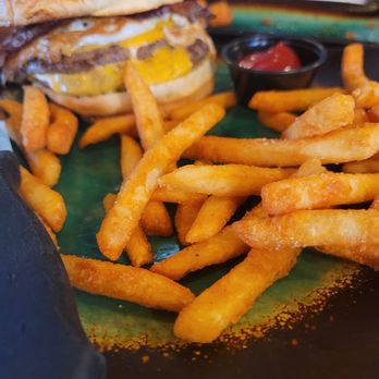 More Brewing - More burger with bacon - A burger with bacon, fried egg, and a bun is served on a tray with a side of French fries. The fries are golden and crispy, with a small container of ketchup nearby. The background shows a dining setting with glasses and utensils. - Brewpubs Near Me - Villa Park, Illinois