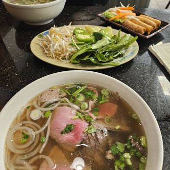 Pho 79 - A bowl of Vietnamese pho featuring sliced beef, rice noodles, green onions, and herbs in a clear broth is placed on a black table. In the background, there is another bowl of pho, a plate with bean sprouts, sliced cucumbers, and leafy greens, and a plate of spring rolls with vegetables. The setting appears to be a casual Vietnamese restaurant with a dark interior and outdoor seating visible. - Vietnamese Near Me - Miami, Florida