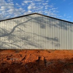 Mitchell Landscaping & Construction - A completed metal storage building features white vertical siding, dark trim, a peaked roof, and freshly graded red soil unde...