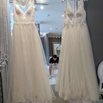Winnie Couture - Two wedding dresses with intricate lace and beadwork on the bodices are hanging on white hangers in front of a large, ornate silver mirror. The dresses feature flowing skirts made of tulle or similar fabric. A person is visible in the background, crouching and looking at their phone, in a room with ceiling lights and a second mirror. The setting appears to be a bridal boutique or fitting room. - Bridal Near Me - Boston, Massachusetts