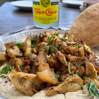 Rumi Middle Eastern Grill - Chicken shawarma hummus plate - A close-up image of a chicken shawarma hummus plate featuring chopped, seasoned chicken on a bed of hummus, garnished with fresh herbs. A piece of pita bread is visible on the side, and a bottle of Topo Chico mineral water is in the background on a wooden table. The setting appears to be a casual dining environment. - Mediterranean Near Me - Chicago, Illinois