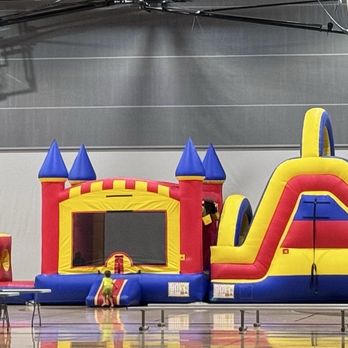 Adventure Commons - Adventure Inflatable Pro Inflatable 18' slide, bounce house & obstacle course - A large inflatable bounce house, castle, and slide setup is arranged inside a gymnasium with a polished wooden floor. The inflatables are brightly colored in yellow, red, and blue, and are positioned side by side. The gym has a high ceiling with visible basketball hoops and sports equipment hanging from above. - Amateur Sports Teams Near Me - Bourbonnais, Illinois