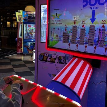 ENTERRIUM - A large arcade game features a colorful screen displaying ticket instructions, with a red-and-white striped cannon and a person operating a control panel. - New American Near Me - Schaumburg, Illinois