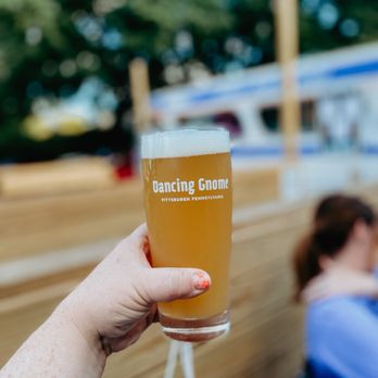 Dancing Gnome - Outside area - A hand with painted nails holds a tall beer glass featuring the 'Dancing Gnome' logo, with an outdoor setting in the background. The background includes blurred trees, a vehicle, and wooden structures, indicating an outdoor brewery or beer garden environment. - Breweries Near Me - Pittsburgh, Pennsylvania