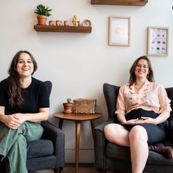 Cosmos Midwifery - Midwives Jenna and Chloë sitting in matching black arm chairs, smiling at the camera.