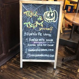 A chalkboard menu with Halloween decorations, including a pumpkin and spider web, listing pumpkin-flavored coffee drinks.