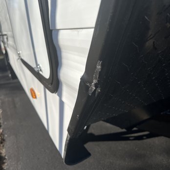 Camping World - Uxbridge - I found this after the unit was basically dropped in my driveway - The image depicts a close-up of the lower side of a white RV or trailer, focusing on a black trim or bumper. The trim shows signs of damage, including a visible crack or break. The background includes a section of asphalt pavement, indicating the vehicle is parked outdoors. The photo emphasizes the damaged area on the black trim, with the white body of the RV or trailer visible in the background. - RV Dealers Near Me - Uxbridge, Massachusetts