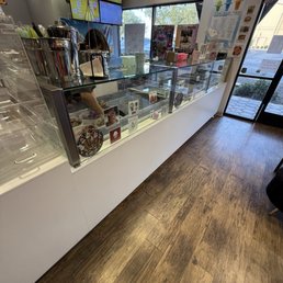 Counter - A glass display case contains multiple sections with different ice cream flavors and Filipino desserts at Scooptopia in Phoenix, AZ. Behind the counter, a staff member is partially visible, with a digital menu screen mounted on the wall above. The shop has a window showing outdoor scenery and posters on the wall near the entrance. The floor is wood, and the overall setting is a casual dessert shop.