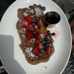 French Toast - French toast with fresh strawberries and blueberries, dusted with powdered sugar, accompanied by a small container of syrup.
