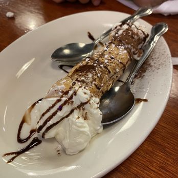 Vinny's Ristorante - Homemade Cannoli - A cannoli dessert featuring a crispy pastry shell filled with creamy filling, topped with whipped cream, chocolate drizzle, and a dusting of powdered sugar. The dessert is served on a white oval plate with two metal spoons placed alongside. The background shows a wooden table and part of a person's hands. - Italian Near Me - Somerville, Massachusetts