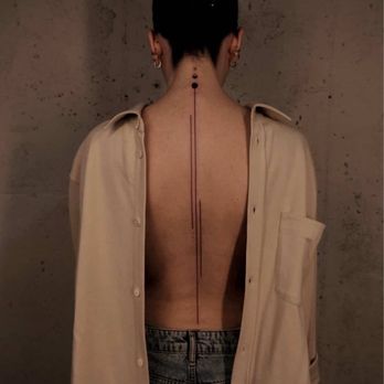 Cafe MSMN - A person with their back to the camera, displaying a vertical tattoo along the spine, wearing an open beige shirt and jeans. The background is a plain, slightly textured wall, and the person's hair is styled in a bun. The tattoo consists of a series of dots and lines running down the center of the back. - Coffee & Tea Near Me - Brooklyn, New York