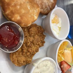 Chicken Fried Chicken - A breakfast meal includes a piece of fried chicken, two biscuits, two cups of eggs (one with a runny yolk), fresh sliced strawberries and melon, and three small containers of condiments: syrup, gravy, and a creamy sauce. The food is arranged on a white plate, with the biscuits and fried chicken taking up most of the space, and the eggs and fruit in small cups.