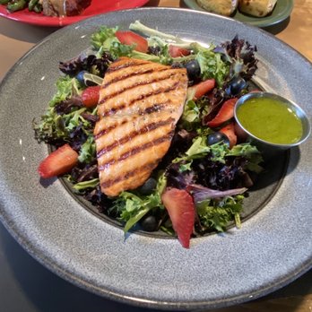 Range Cafe - Grilled Salmon Berry - A plate features a grilled salmon fillet with grill marks, resting on a bed of mixed greens including lettuce, spinach, and arugula, garnished with sliced strawberries and blueberries. A small metal cup contains a green dressing or sauce. In the background, there are additional dishes including a plate with meat and green beans, and a small green plate with two bread rolls. The setting appears to be a casual dining restaurant. - Breakfast & Brunch Near Me - Albuquerque, New Mexico