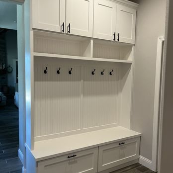 Lawrence Construction Services - A white hall tree features upper cabinets with black handles, open shelving, and six black hooks for hanging items. Below, there is a bench with two drawers for storage. The unit is positioned against a wall in a home entryway, with wood-look flooring and a neutral wall color. - Decks & Railing Near Me - Azle, Texas