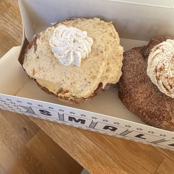 Parlor Doughnuts - Pumpkin Chai Cheesecake Layered Doughnut - The image shows two large donuts inside a bakery box placed on a wooden table. One donut appears to be a vanilla or plain flavor with a dollop of whipped cream on top, while the other is a chocolate or cinnamon-flavored donut also topped with whipped cream. The box has a printed design on its side, and part of a person's clothing is visible at the bottom of the image. - Donuts Near Me - Denver, Colorado