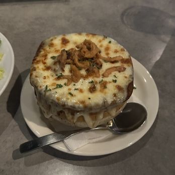 Ithaca Ale House - Six onion soup! - A bowl of six onion soup, topped with melted cheese and fried onions, is served on a white plate with a spoon resting on a napkin. The soup is presented in a round bowl, and the setting appears to be in a restaurant with a gray table surface. In the background, part of another dish and a person wearing a blue shirt are visible. - New American Near Me - Ithaca, New York
