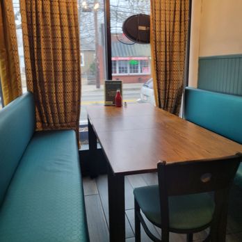Grainsley's Kitchen + Drinks - The image shows a corner booth with teal upholstered seating along two sides of a wooden table inside a restaurant. A single chair with a matching teal seat is positioned at the end of the table. Behind the booth, there is a large window with patterned curtains, revealing a street scene outside. A small menu and a condiment bottle are placed on the table, and a hanging light fixture is visible above. The setting appears to be a casual dining area. - Cocktail Bars Near Me - Warwick, Rhode Island