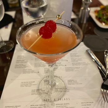 Circa69 - Drinks - A pinkish cocktail with a cherry garnish on a skewer is on a restaurant table, with a menu, water glass, and other tableware visible. - Cocktail Bars Near Me - Jensen Beach, Florida