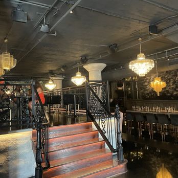 Dearly Beloved - The image shows a restaurant interior with a staircase leading to an upper level, chandeliers hanging from the ceiling, and a bar area with seating. - Cocktail Bars Near Me - Chicago, Illinois