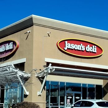 Jason's Deli - Jason's Deli - The image depicts the storefront of Jason's Deli in Albuquerque, NM. The building has a beige exterior with two prominent signs displaying the restaurant's name. The signage is oval-shaped with a red background and white text. The entrance features glass doors and windows, with a white awning extending over the sidewalk. A white car is parked in front of the store, and a leafless tree is visible on the left side of the image. The sky is clear and blue. - Delis Near Me - Albuquerque, New Mexico
