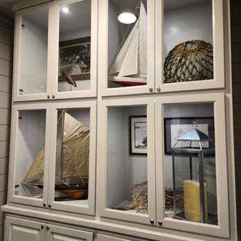 Lindy's Landing - A white display cabinet with glass doors contains model sailboats, a decorative lantern, and framed pictures, set against a wall with wood paneling. - American Near Me - Wauconda, Illinois