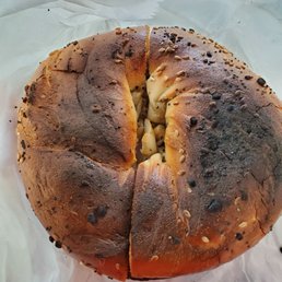 I ordered an everything bagel toasted with butter and this is what I got, didn't say anything and passed it off like this. Horrible - A round, toasted bagel with a dark, crispy crust is shown, topped with various seeds and spices. The bagel appears to be slightly cracked and unevenly toasted, with some areas darker than others. It is placed on white paper, which is crinkled and slightly translucent. The interior of the bagel is partially visible through a crack, showing a soft, dense texture.