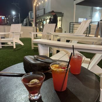 The Station - Outdoor patio scene at night featuring white Adirondack chairs arranged around a black circular table. On the table are three cocktails with straws and garnishes, and a black purse. String lights hang overhead, and a building with a window and a person standing nearby are visible in the background. - Pizza Near Me - Jackson, Mississippi