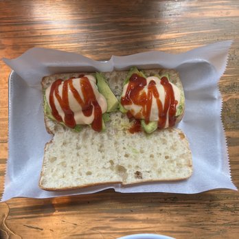 Maya Congee Cafe - Breakfast sandwich - A sandwich featuring two eggs topped with ketchup and mayonnaise, along with slices of avocado, is served on a piece of bread. The sandwich is placed on a paper-lined tray on a wooden table. Part of a bowl with greens and avocado is visible at the bottom of the image. The setting appears to be a casual dining environment. - Sandwiches Near Me - Brooklyn, New York