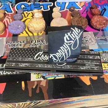 Cookie Plug - A variety of cookies and snack items are arranged on a colorful table at Cookie Plug, including different flavors and packaging, with promotional signs. - Desserts Near Me - Avondale, Arizona