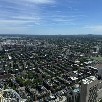 View Boston - A high-angle view of a city with numerous buildings, streets, and green spaces under a blue sky with scattered clouds. - Observatories Near Me - Boston, Massachusetts