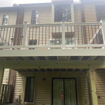 Montgomery Decks - The image shows a newly built balcony with white railing and black vertical balusters attached to a beige multi-story residential building. The balcony is supported by wooden beams and posts, with a sliding glass door leading to the interior. The building has multiple windows, and the construction appears recent, with visible wood framing and no finishing touches yet. - Decks & Railing Near Me - Frederick, Maryland