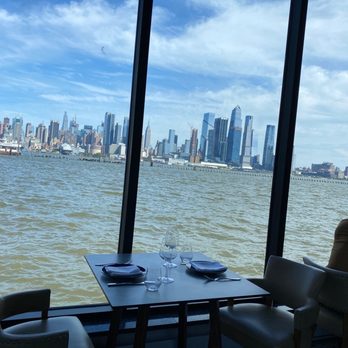 Molos - A dining table set for two with wine glasses, plates, and napkins is placed near large floor-to-ceiling windows. The windows offer a panoramic view of the New York City skyline across the water, with tall skyscrapers visible under a partly cloudy sky. The interior includes chairs with armrests, and part of a person is visible on the right side of the image. - Greek Near Me - Weehawken, New Jersey