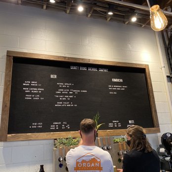 Gravity Bound Brewing - Menu - Two people are seated at a bar in front of a large black menu board framed with wood, listing beer, kombucha, and Wi-Fi options at Gravity Bound Brewing in Albuquerque. One person has short hair and a white shirt with an orange logo, while the other has long hair and wears a black mask. The setting features a white brick wall, a visible light bulb, and a coffee machine on the right. - Brewpubs Near Me - Albuquerque, New Mexico