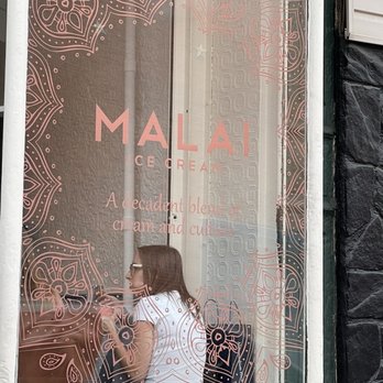 Malai - A woman with glasses and long hair is sitting inside Malai ice cream shop, visible through a window with pink decorative patterns and text. - Ice Cream & Frozen Yogurt Near Me - Brooklyn, New York