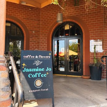 Jasmine Jo - Jasmine Jo - The image shows the entrance of Jasmine Jo Coffee, a coffee and flower shop located in Tempe, AZ. The storefront has a brick facade with two arched windows and a glass door. A black signboard placed on the sidewalk displays the shop's name, 'Jasmine Jo Coffee,' along with the words 'coffee & flowers' and 'support local.' The sign also features a coffee cup graphic and decorative stars. There are outdoor seating areas visible through the glass door, with umbrellas and tables outside. A large potted plant is positioned near the entrance, and hanging plants are seen above the doorway. - Coffee & Tea Near Me - Tempe, Arizona