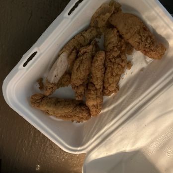 Fat Shack - The image shows several fried chicken tenders inside a white foam takeout container. The tenders have a crispy, golden-brown coating and are arranged randomly. The container is open, revealing the food, and is placed on a dark surface. The photo appears to be taken indoors or in a shaded area, with a focus on the fried chicken as the main subject. - Burgers Near Me - Manhattan, Kansas