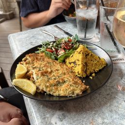 Parmesan crusted whitefish - A black plate holds a breaded whitefish fillet garnished with lemon wedges, accompanied by yellow rice with black rice grains and a mixed green salad with cherry tomatoes. The meal is set on a table with utensils, glasses of water, and a glass of white wine. The setting appears to be an outdoor restaurant with patio seating, and a person in a dark shirt is visible in the background, holding a bowl.