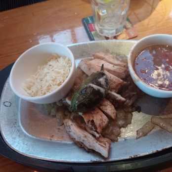 Silverado - Fajitas - A metal platter holds fajitas with grilled meat, sliced and topped with a jalapeño pepper. Accompanying sides include a small bowl of rice and a bowl of salsa. A glass of water is visible in the background on a wooden table. - American Near Me - Annandale, Virginia
