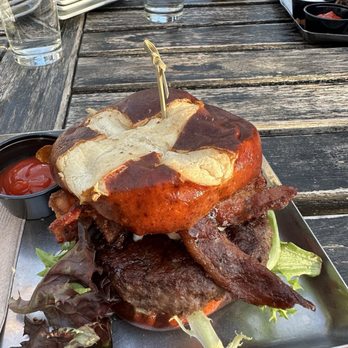 Empire - G.O.A.T burger - A sizable burger featuring a toasted bun, crispy bacon, and fresh lettuce is presented on a metal tray. The burger is topped with a large, golden-brown bun and garnished with a small skewer. Surrounding the burger are small portions of ketchup and leafy greens. The setting includes an outdoor wooden table with glasses of water, a beer, and other condiments, indicating a casual dining environment. - Burgers Near Me - Naperville, Illinois