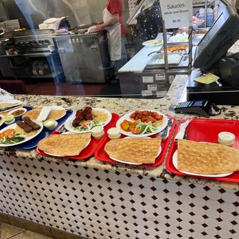 Flame Kabob - a variety of food items on a counter - Several trays of Afghan dishes, such as naan bread, rice, and various meat dishes, are arranged on a granite counter at a restaurant. Small containers of sauce accompany each tray. The background shows a kitchen area with staff preparing food and a sign indicating sauces priced at $0.35. The counter has a point-of-sale system, a cash register, and a cup with pens. - Afghan Near Me - Springfield, Virginia
